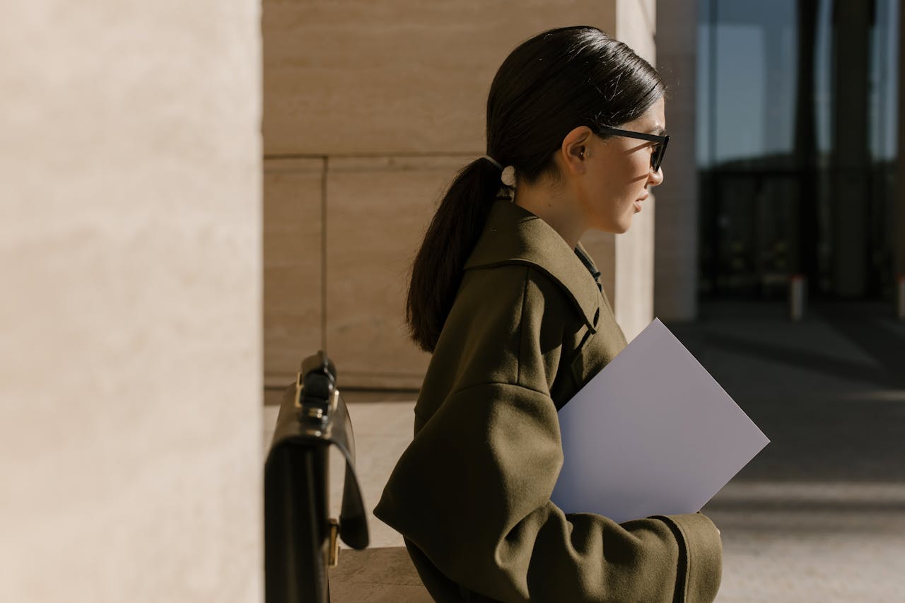 Side view of a woman in an olive coat holding a document, standing outdoors.